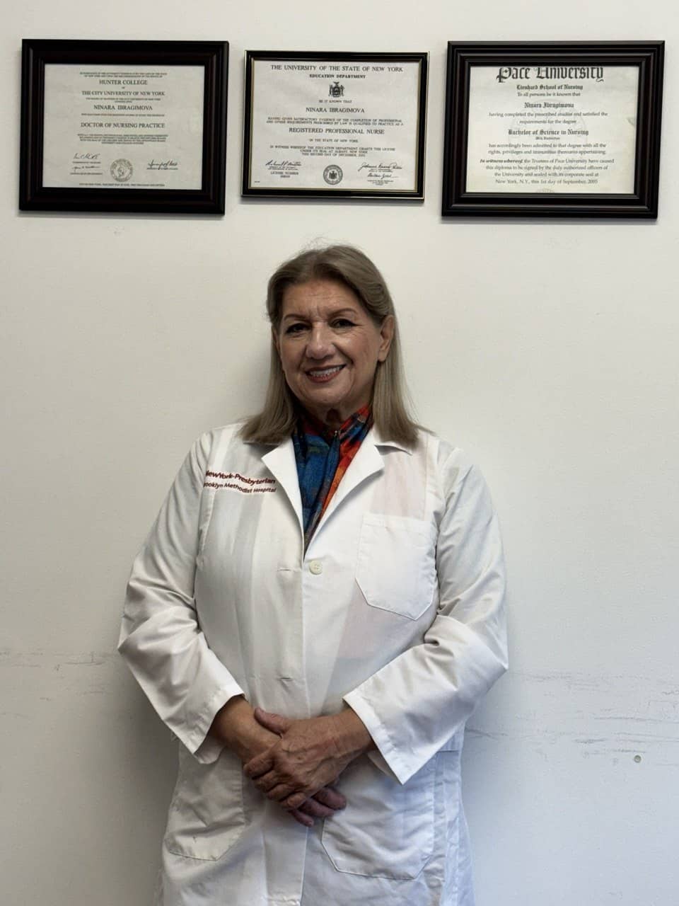 Ninara Ibragimova in a white coat smiling in front of framed Hunter College, New York State and Pace University diplomas